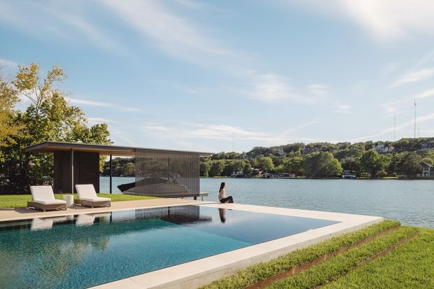 A contemporary waterfront outdoor area with a long rectangular swimming pool extending toward a lake or river. The pool terrace includes pale paving, two lounge chairs, and a slim modern pavilion with a flat roof and dark screened walls. A person sits at the water's edge, with landscaped lawn and wooded residential hills visible across the water.