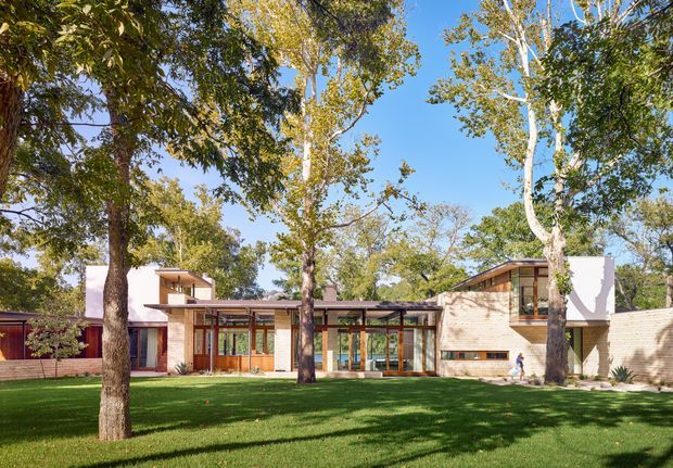 Wide exterior view of a low-slung modern residence set within a mature, landscaped garden. The house features warm brick masonry, extensive timber-framed glazing, flat and shallow-pitched roof forms, white rendered wall elements, and a long glazed connection between wings. Large established trees and a broad lawn dominate the foreground.