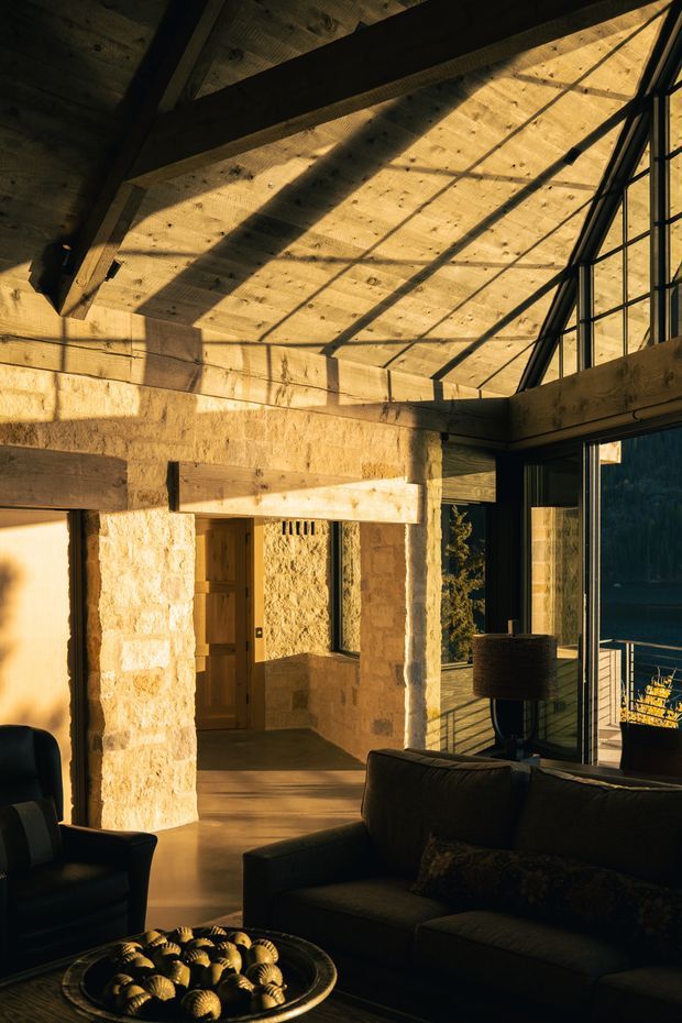 A warm, dramatic interior living space with exposed heavy timber roof beams and ceiling boards, thick rough-cut pale stone walls, and expansive black-framed glazing opening toward a deck and natural landscape. A dark sofa, armchair, and low coffee table occupy the foreground, while strong late-afternoon sunlight casts bold geometric shadows across the room. The space has a rustic lodge-like character with a high pitched roof and generous glazed opening.
