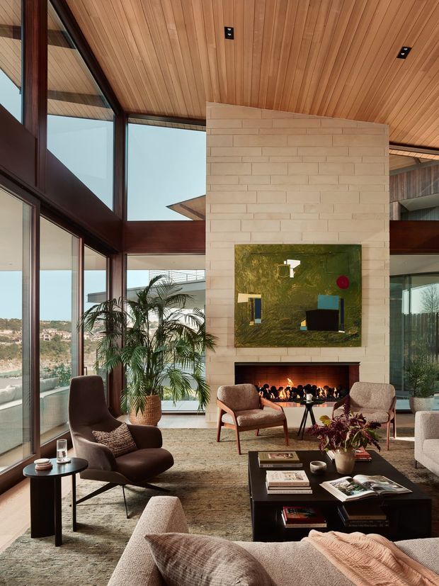 A spacious, high-ceilinged living room with floor-to-ceiling glazing, dark timber-framed windows, and a warm timber-lined ceiling. A pale stacked-brick fireplace wall with a contemporary open fire anchors the room, with abstract artwork above it. The room is furnished with mid-century-inspired lounge chairs, sofas, a black coffee table, side tables, rugs, and indoor palms. A hillside landscape is visible through the glazing.