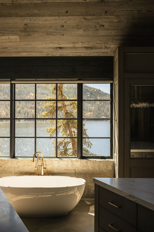 A warm, moody bathroom interior with a freestanding white bathtub positioned beside large black-framed grid windows overlooking a lake and wooded hills. The room features dark wood wall and ceiling panelling, a textured beige plaster wall beneath the windows, dark built-in cabinetry with brass pulls, and a pale stone vanity benchtop. Natural light creates strong reflections across the tub and floor.
