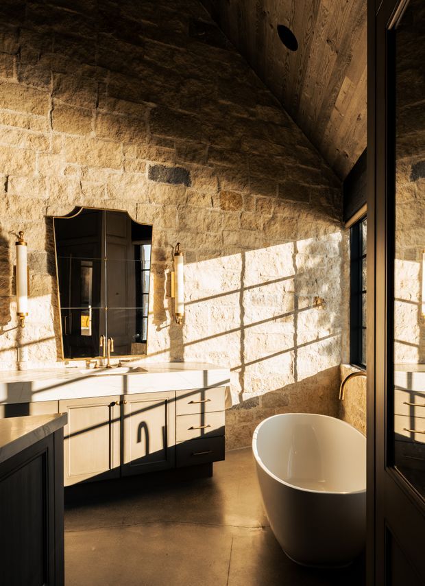 A warmly lit, high-end bathroom with exposed rough-cut stone masonry walls, a freestanding white oval bathtub, and a floating timber-toned vanity with dark framed cabinetry and brass fittings. A large black-framed mirror and glazing reflect the room, while strong late-afternoon sunlight creates geometric shadows across the stone and concrete floor. The space has a rustic yet refined, dramatic character.