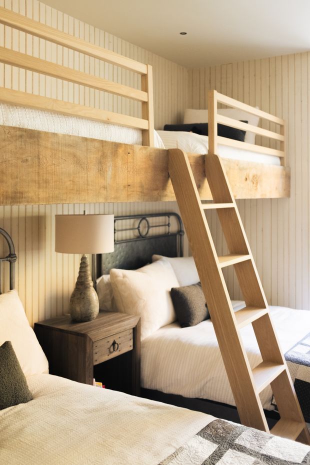 A warm, compact bunk bedroom with multiple built-in timber bunk beds, a wooden ladder, white bedding, and a dark wood bedside table with a lamp. The walls are clad in light vertical timber boards, creating a cozy cabin-like atmosphere.