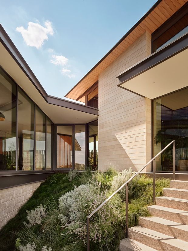 Exterior view of a contemporary low-rise residence organised around a landscaped courtyard. The composition shows pale horizontal masonry, extensive dark-framed glazing, warm timber soffits and cladding, projecting rooflines, and a stair with a slim metal handrail leading to a recessed entrance. Ornamental grasses and silvery planting fill the courtyard garden under a bright sky.