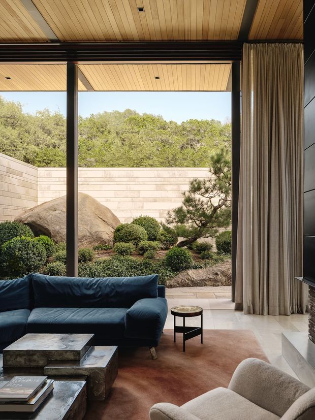 A refined living room with a deep blue sectional sofa and sculptural pale armchair arranged around low stone coffee tables. Full-height dark-framed glazing opens to a landscaped courtyard with rocks, clipped shrubs and a small tree. The room features a warm timber-lined ceiling, floor-to-ceiling curtains, pale stone flooring, a dark tiled fireplace wall, and a large muted rug. The composition is calm and sophisticated, with strong indoor-outdoor connection and natural materials.