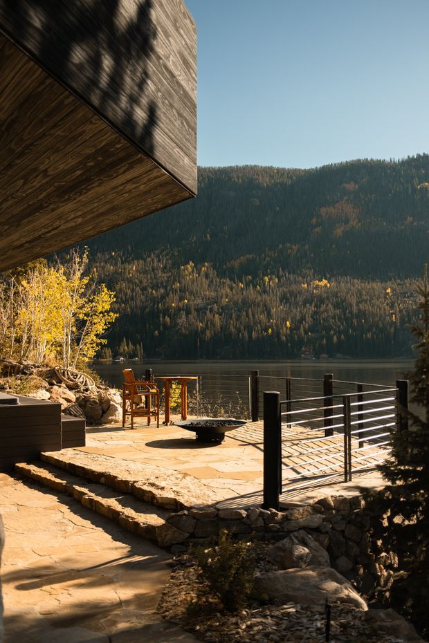 A lakeside outdoor terrace beneath a projecting timber-clad roof or upper-level volume. The terrace is paved with irregular natural stone and bordered by rugged stacked-stone retaining walls, dark horizontal-planter joinery, and black metal posts with horizontal cable or bar railing. A central low fire bowl and outdoor seating create a relaxed entertaining area overlooking a forested mountain lake. Warm sunlight and autumn foliage give the scene a natural, tranquil atmosphere.
