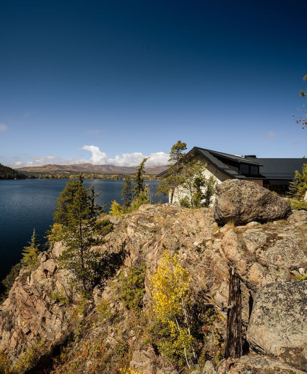Exterior view of a dark-roofed house set into a rugged rocky shoreline above a calm lake, with forested and mountainous terrain across the water. The building has a steeply pitched roof, dark metal roofing and dark-framed glazing, with light-coloured exterior walls partially visible among natural rock outcrops and sparse trees. The image is a bright, scenic architectural landscape photograph.