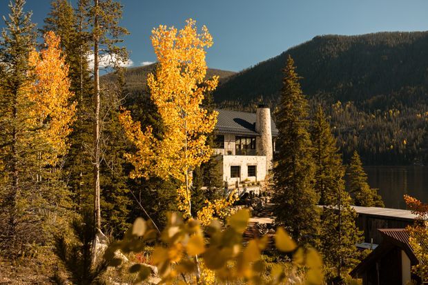A multi-level mountain home partially screened by golden autumn trees, set among dense coniferous forest beside a calm lake. The building has a light masonry facade, dark pitched metal roof, substantial stone chimney, and extensive glazing, with additional dark-roofed structures visible in the foreground.