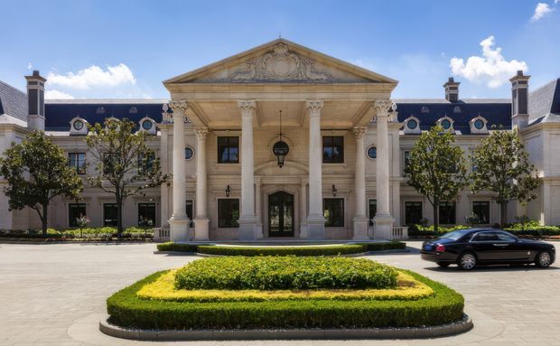 Formal grand architectural exterior featuring a symmetrical classical facade with a prominent portico, tall fluted columns, ornate capitals, triangular pediment, sculptural detailing, dormer windows, and dark slate roofing. Manicured hedges and a circular driveway frame the entrance.