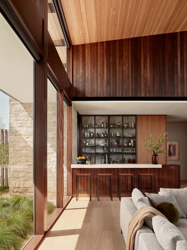 A warm contemporary interior combining a living area and a built-in home bar. Full-height timber-framed glazing opens toward a landscaped exterior, while rich vertical timber wall panelling and a timber-lined ceiling create a cohesive architectural feature. The bar has dark glazed display cabinetry, a light stone or composite benchtop, fluted timber cabinetry, and four slim bar stools. A large light upholstered sectional sofa occupies the foreground.