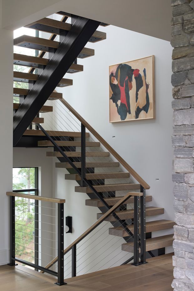 A bright contemporary stairway with open-riser timber treads, black steel stringers and posts, and horizontal cable balustrades. The stair rises through a multi-level void beside large windows and a skylight. A rough stacked-stone wall frames the right side, while a colourful abstract artwork hangs on the pale wall.