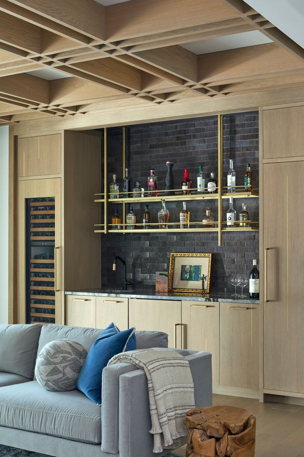 A refined living room with a built-in home bar and integrated wine fridge. The bar features light timber cabinetry, a dark textured tile backsplash, brass-toned open shelving, a stone benchtop, sink and concealed storage. A sculptural exposed timber coffered ceiling adds strong architectural character. In the foreground is a soft grey sofa with blue and patterned cushions, a draped throw, and a carved timber side table.