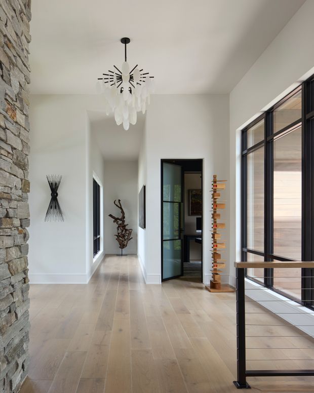 Bright contemporary residential hallway or entry landing with pale wide-plank timber flooring, white walls and ceiling, a dramatic sculptural pendant light, and a black metal-framed glazed opening overlooking a stairwell. A rough stacked-stone feature wall lines the left side, while timber and sculptural art pieces add visual interest. The space connects to a darker room through a black-framed glass door and includes a black metal stair balustrade.