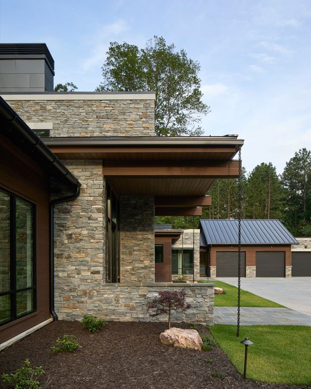 Exterior view of a contemporary multi-level residence with extensive irregular stacked-stone wall cladding, warm timber soffits, dark metal roof edges, and large glazed openings. A covered outdoor transition and landscaped garden lead toward a detached-looking garage volume with a pitched standing-seam metal roof and stone-and-timber finishes. The setting includes mature trees and a paved driveway.