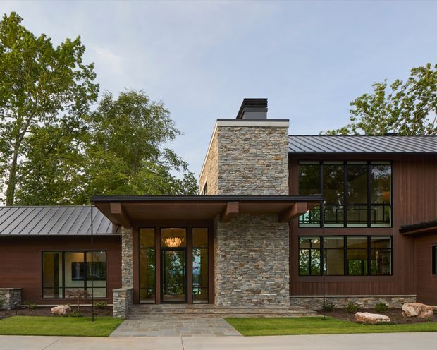 Front exterior view of a modern multi-volume house set among mature trees. The facade combines horizontally layered natural stone masonry, dark vertical timber cladding, extensive black-framed glazing, and low-pitched standing-seam metal roofs. A projecting timber-and-metal entry canopy leads to glazed double entry doors, while landscaped lawn and stone paving frame the approach.