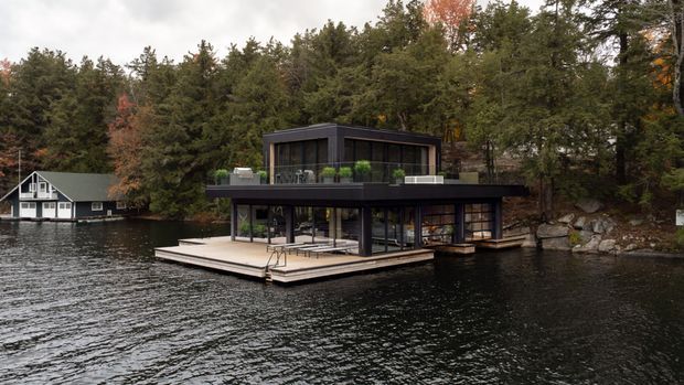 Exterior view of a contemporary two-storey lakeside house extending over the water, with dark cladding, extensive floor-to-ceiling glazing, broad flat rooflines, and a large timber deck. Dense forest, rocky shoreline, and a neighbouring waterside home form the background.