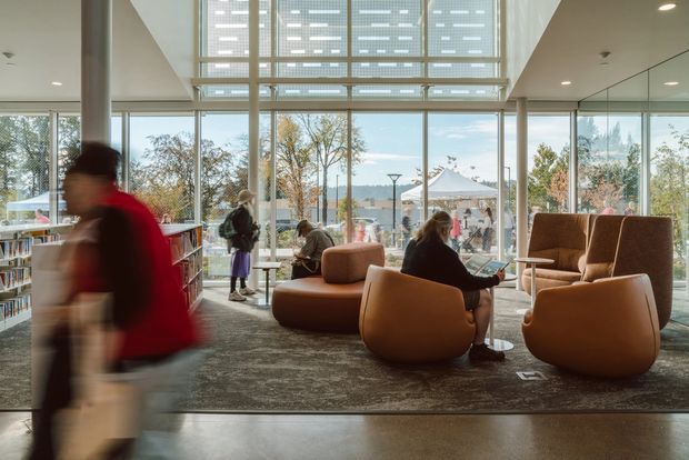 A bright, contemporary library or public reading lounge with floor-to-ceiling glazing, a large overhead clerestory/skylight structure, bookshelves along the left wall, and clustered sculptural brown lounge chairs around low tables. People are using the space, with trees and an outdoor courtyard visible beyond the glazing. The interior has a spacious, airy civic or commercial character.