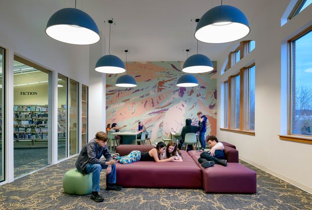 A bright contemporary library lounge and reading area with large windows, glass partitions, suspended dome pendant lights, a colourful abstract mural, and a cluster of low burgundy modular sofas and ottomans. Several people are reading or using the communal tables. Bookshelves and a sign reading “FICTION” are visible through the glazed partition on the left. The space has patterned carpet, warm timber window frames, and an airy open layout.