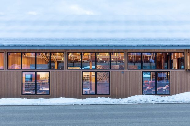 A long, low modern building facade viewed from across a road in a snowy setting. The exterior is clad in vertical brown timber boards and features extensive dark-framed glazing, with warm-lit interior spaces and exposed timber structure visible through the windows. Snow rests along the base and roof edge, creating a cool blue-and-warm-amber contrast.