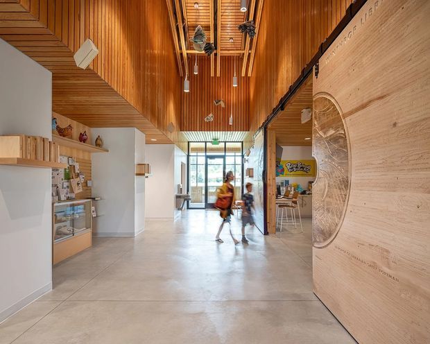 A bright, spacious multi-purpose interior with a long polished concrete floor and dramatic high timber-lined ceiling. The central circulation space leads toward full-height glazing at the rear, with built-in timber cabinetry and display shelving on the left, a timber-clad feature wall and circular relief detail on the right, and an open activity or communal area beyond. Hanging pendant lights and exposed timber ceiling battens add warmth and visual interest. Blurred people are present in the hallway.
