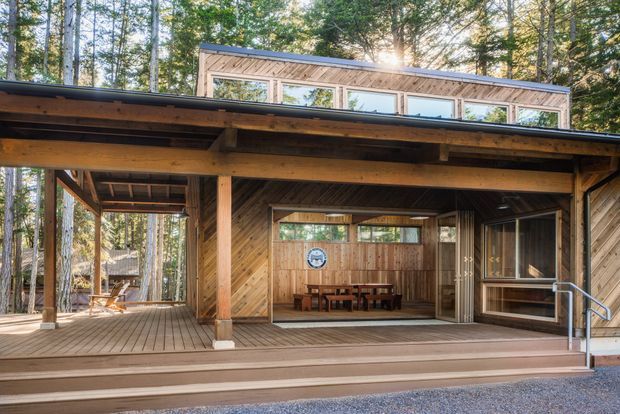 Exterior view of a timber-clad, forest-set house with a broad covered deck and open outdoor dining or living area. The building has a low-pitched roof, exposed timber beams and posts, diagonal wood siding, extensive glazing, and an upper clerestory-like window band. Steps lead up from a gravel forecourt to the deck, with a handrail at the right.