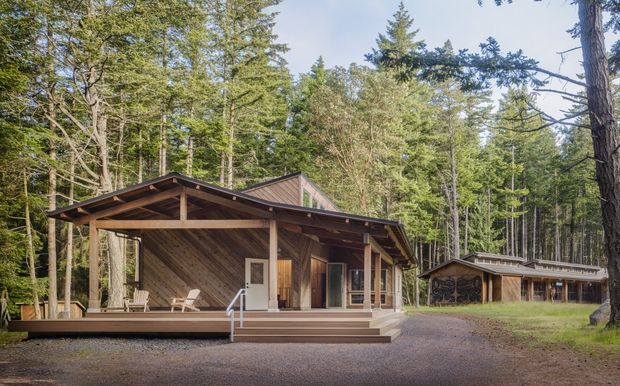 A single-storey timber cabin or lodge set within a dense forest. The building has a broad gabled roof, deep covered front veranda, exposed timber framing and diagonal wood wall cladding. A detached timber outbuilding is visible to the right, connected by a gravel driveway or forecourt. The scene has a rustic woodland character with natural timber tones and muted green surroundings.