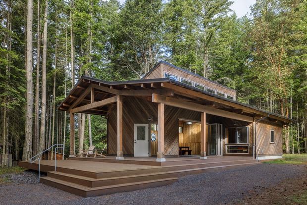 A rustic timber-clad house or cabin set within a dense forest. The single-storey building features a broad covered veranda, exposed timber posts and rafters, a raised timber deck with steps and accessible handrails, large glazed openings, and a pitched gable roof with a long clerestory-like upper window band. The surrounding woodland and gravel foreground reinforce its secluded natural setting.