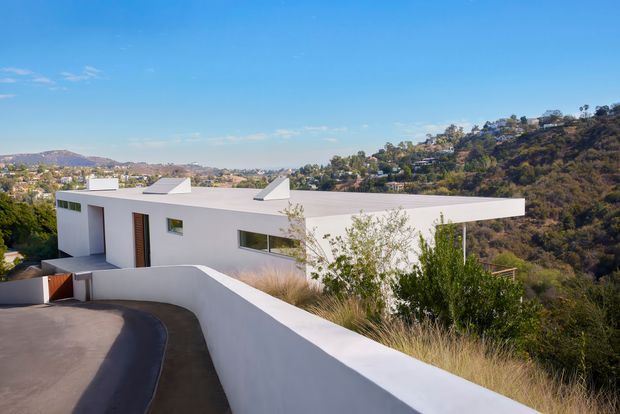 A modern white flat-roofed house set on a steep, elevated suburban hillside. The angular building has clean rectangular volumes, projecting roof planes, narrow horizontal windows, and dark timber-toned entry detailing. A curved driveway and white boundary wall lead toward the residence, with wooded hills and neighbouring homes visible beyond.