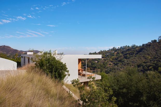 Exterior view of a modern white flat-roofed house set into a steep, wooded hillside. The building has clean geometric volumes, broad projecting roof planes, large openings, and a timber-toned balcony or terrace overlooking the surrounding landscape. Dry ornamental grasses and mature vegetation frame the foreground, with hills visible beyond under a clear blue sky.