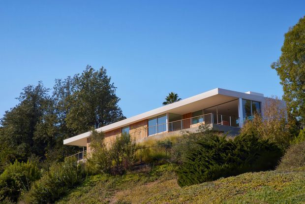 Exterior view of a modern, low-profile flat-roof residence set into a landscaped, sloping site. The house features broad horizontal roof planes, extensive glazing, timber-toned wall surfaces, and open upper-level balconies with slender metal balustrades, surrounded by mature vegetation under a clear blue sky.