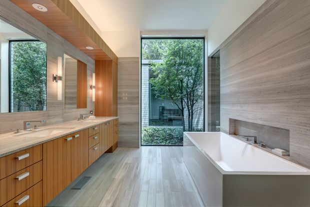 A spacious contemporary bathroom with a double vanity in warm vertical timber cabinetry, pale stone benchtop, large mirrors, a freestanding white bathtub, and extensive grey stone-look wall and floor finishes. Full-height glazing opens the room visually to a leafy courtyard, while recessed ceiling lights and wall sconces provide layered illumination.