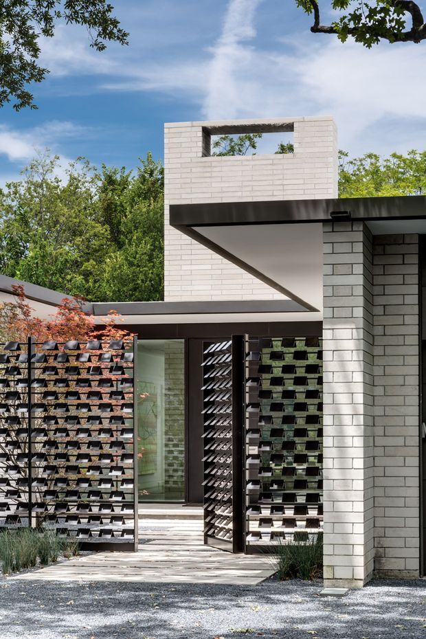 A contemporary residential facade framed by mature landscaping, featuring pale brick masonry, crisp flat rooflines, deep black metal eaves, and a sculptural upper-level opening. A central glazed entry is screened by distinctive black geometric gates and planted trellis panels, with a paved path leading through the landscaped forecourt.