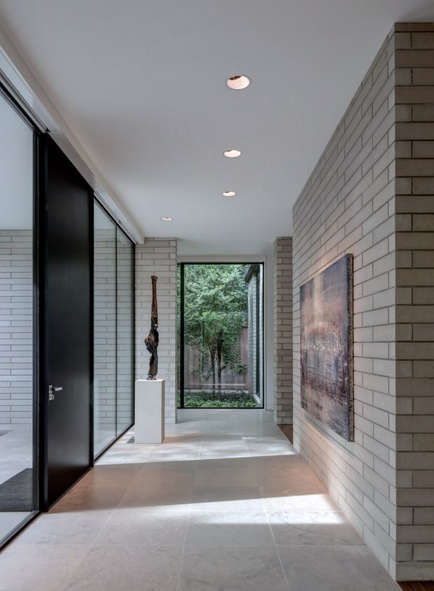 A refined residential entry hall with a long, spacious circulation path. Pale masonry brick walls line the right side and frame the rear opening, while a large black-framed glazed door and full-height glazing run along the left. Large light stone-look floor tiles, a smooth white ceiling with recessed downlights, a sculptural object on a white pedestal, and a large wall artwork create a minimalist, gallery-like atmosphere. A leafy courtyard is visible through the rear window.