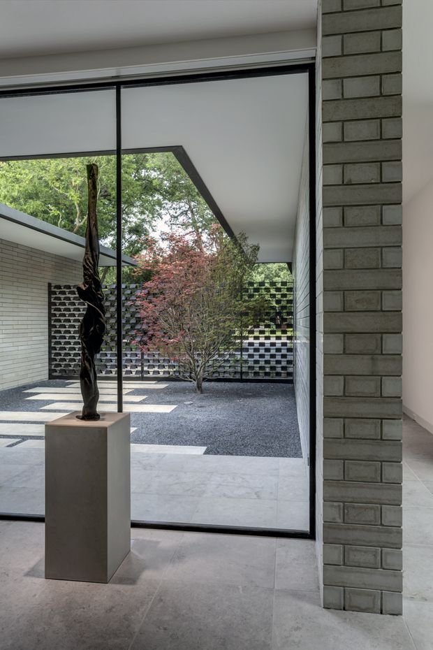A refined modern interior looking through full-height glazing into a sheltered courtyard. The space features large pale stone or concrete-look floor tiles, exposed grey brick walls, slim black-framed glazing, and a minimalist sculptural artwork on a square pedestal. Outside, a gravel courtyard contains a small tree and patterned breeze-block masonry screens beneath a projecting roof, creating strong geometric shadows and a calm indoor–outdoor connection.