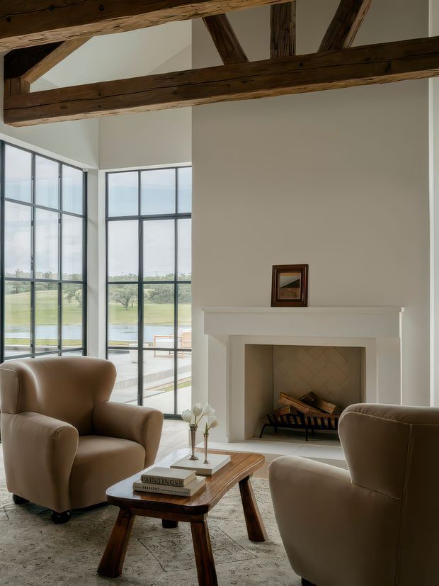 A refined living room with a white painted fireplace surround, herringbone-lined firebox and wood-burning fireplace, framed by expansive black-framed floor-to-ceiling windows overlooking a landscaped property. Exposed dark timber roof trusses contrast with the pale walls and ceiling. Two rounded taupe upholstered armchairs face a low timber coffee table with books and a small floral arrangement, over a large patterned rug.