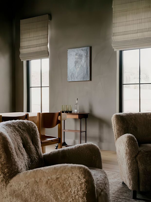 A softly lit living room with two tall black-framed windows fitted with woven Roman blinds, warm taupe plaster walls, and a compact wood console table beneath a framed artwork. Several rounded, textured upholstered armchairs in beige and brown occupy the foreground, with a small dining or writing chair and table visible near the window. The room has a warm, refined, contemporary feel.