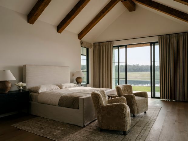 A refined bedroom with a low-profile upholstered bed, bedside tables and lamps, two textured lounge chairs, and a large patterned rug. Full-height glazed doors and windows open to a calm waterside landscape, while exposed dark timber roof beams and a vaulted ceiling add warmth and architectural character. Neutral cream, beige and brown tones create a serene, luxurious atmosphere.