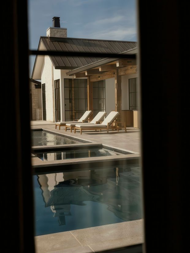 A refined residential pool courtyard viewed through a dark-framed opening. The scene features a pale rendered house with a dark standing-seam metal roof, timber-framed covered veranda, tall black-framed glazed doors, and several light lounge chairs beside a tiered reflecting pool and swimming pool. The architecture combines clean contemporary lines with warm timber detailing and a relaxed resort-like atmosphere.
