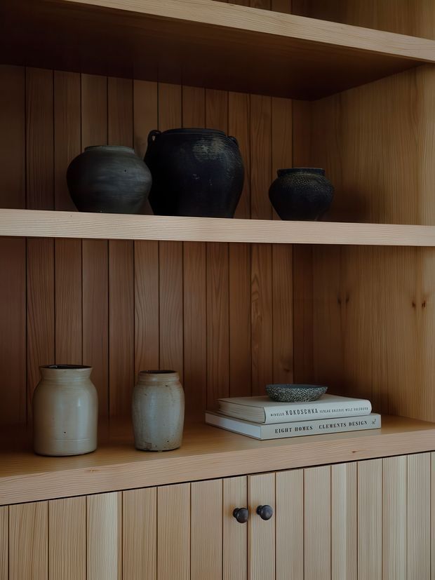 Close-up interior photograph of custom built-in timber shelving and lower cabinetry with vertical wood panelling. The open shelves display dark ceramic vessels, pale pottery, and a small stack of design books, creating a warm, curated storage and display feature.