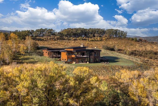 Wide exterior view of a low-profile contemporary house set within an expansive, dry native landscape. The home features dark timber or brown cladding, extensive glazing, projecting flat roof forms, and an integrated deck or terrace. Rolling hills and mature vegetation surround the property under a bright blue sky with clouds.
