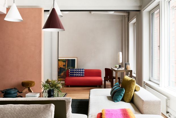 A bright, eclectic living room with large industrial-style windows, a red sofa, neutral textured seating, colourful cushions and a vivid throw. A terracotta-toned feature wall, pendant lights, timber flooring and a compact dining/work area create a warm contemporary interior.