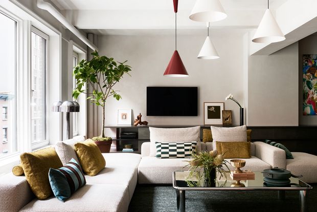 Bright contemporary living room with large windows, a light sectional sofa, patterned cushions, black low-profile media cabinetry, a wall-mounted television, and multiple sculptural pendant lights. The room combines pale textured walls and upholstery with dark timber or painted joinery, metallic accents, indoor planting, and colourful artwork.