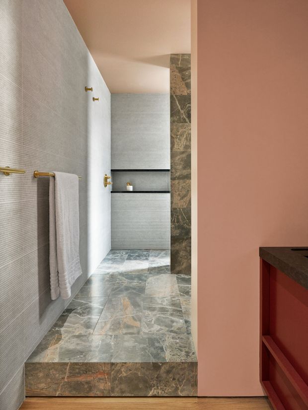 A contemporary bathroom viewed from an adjoining space, featuring a walk-in tiled shower at the rear, grey textured wall tiles, large mottled natural-stone floor and step tiles, dark stone shelving and wall cladding, and brushed brass towel rails and fixtures. A warm dusty-pink wall and red-toned built-in cabinetry are visible in the foreground.