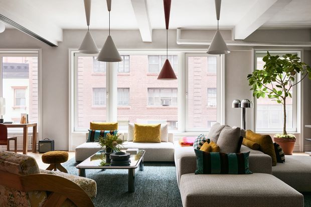 Bright contemporary living room with a large L-shaped light-grey sectional sofa, patterned and velvet accent cushions, a glass coffee table, and a textured blue-grey rug. Multiple sculptural pendant lights hang from a white coffered and beam-detailed ceiling. Large windows overlook adjacent brick buildings, while a tall indoor tree and layered furnishings add warmth and colour.