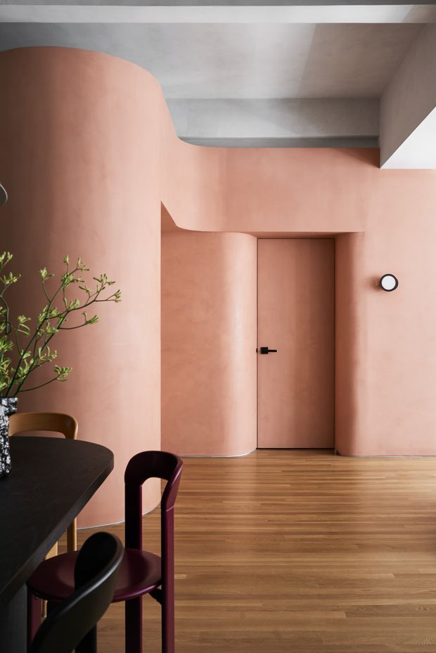 A refined interior dining area with softly curved plastered walls in a warm terracotta-pink tone, a flush matching door, black wall light, and warm timber flooring. A dark dining table with sculptural burgundy and black chairs occupies the foreground, while a small branch arrangement adds organic detail. The ceiling and boxed architectural elements are light grey, creating a restrained contemporary composition with strong rounded forms.