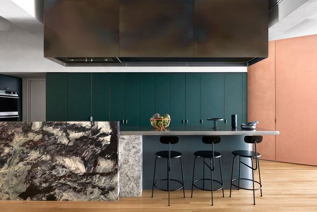 A bold contemporary kitchen centred on a large island with a dramatic dark, veined stone waterfall end and a pale stone benchtop. Deep teal full-height cabinetry lines the rear wall, with a dark overhead rangehood enclosure and integrated appliances. Three black metal bar stools sit along the island, while a terracotta-toned wall and light timber flooring add warmth. The space has a refined, high-contrast palette and strong sculptural materiality.