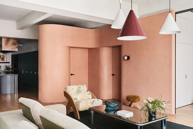 Contemporary open-plan living room with a sculptural terracotta plaster feature wall and curved partition enclosing a doorway. The room has pale upholstered seating, a patterned lounge chair, a black coffee table, pendant lights in white and burgundy, timber flooring, and dark built-in cabinetry in the background.