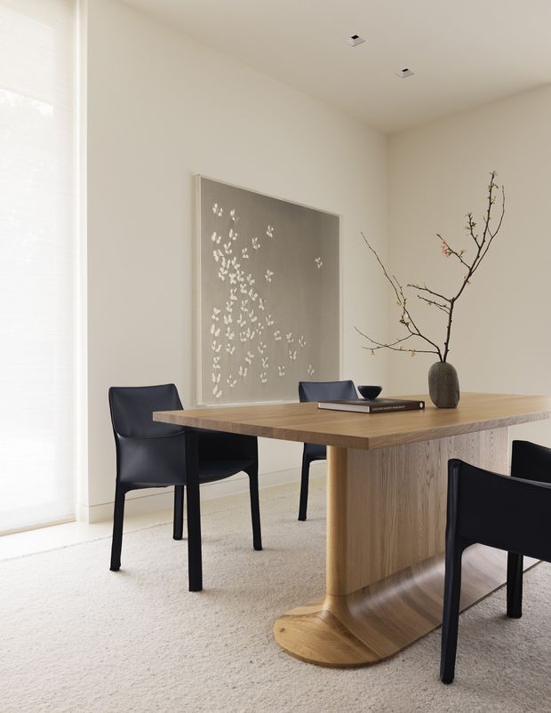 A bright, minimalist dining room with a large light timber dining table featuring a sculptural pedestal base, surrounded by black upholstered dining chairs. The room has warm off-white walls, pale carpet, a full-height window with a translucent blind, recessed ceiling lights, and restrained decor including a monochrome wall artwork, a shallow bowl, books, and a branch arrangement.