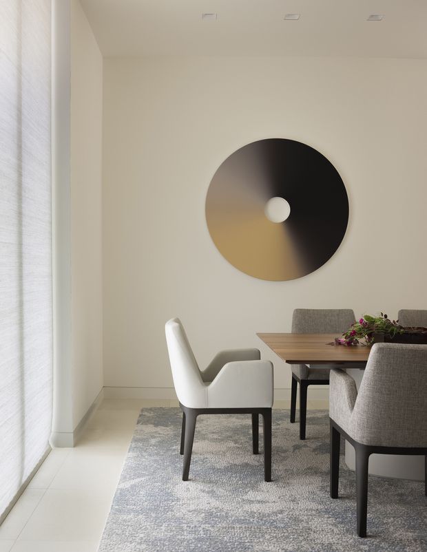 A refined dining area with a rectangular timber dining table and upholstered dining chairs arranged on a large grey patterned rug. The room has warm off-white walls, pale tiled flooring, sheer floor-to-ceiling window coverings, recessed ceiling lights, and a large circular metallic wall feature with a central opening. The restrained palette and clean lines create a calm, contemporary interior.