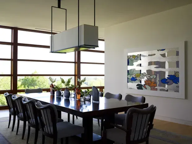 A refined dining room with a large dark rectangular dining table surrounded by upholstered and spindle-back chairs. Floor-to-ceiling timber-framed glazing fills the left side, providing abundant natural light and views of greenery. A contemporary rectangular pendant light hangs above the table, while a colourful abstract artwork provides a feature on the white wall. The room has a warm, sophisticated, modern character.